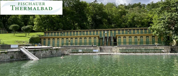 Das Bild zeigt einen Swimmingpool mit einer Rutsche, umgeben von einem großen Gebäude mit grünen Türen. Bäume und ein Rasenfeld sind im Hintergrund zu sehen.