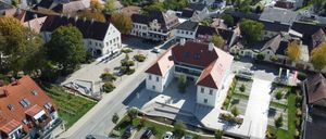 Ein Luftbild einer Stadt zeigt verschiedene Gebäude, darunter ein großes weißes Gebäude mit einem roten Dach. Umgeben sind es von anderen Gebäuden, einige mit roten Dächern. Autos sind auf der Straße geparkt.