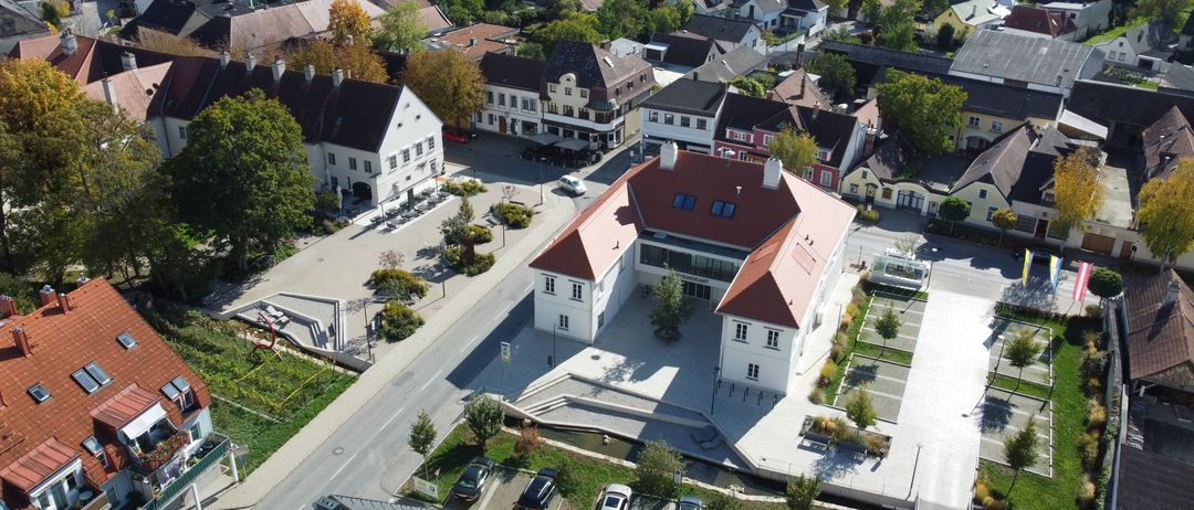 Ein Luftbild einer Stadt zeigt verschiedene Gebäude, darunter ein großes weißes Gebäude mit einem roten Dach. Umgeben sind es von anderen Gebäuden, einige mit roten Dächern. Autos sind auf der Straße geparkt.