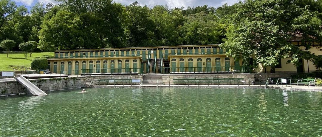 A large pool with a stone wall and a yellow building with many doors and windows. Trees surround the area. A person swims in the pool.