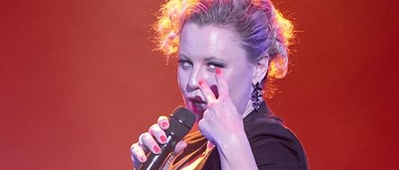 Eine Frau tritt auf der Bühne auf, singt in ein Mikrofon und trägt ein blaues Kleid und eine schwarze Bluse.