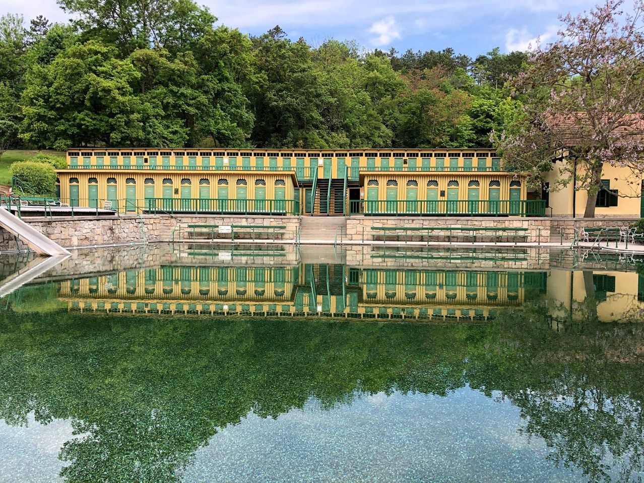 Ein leuchtendes gelbes und grünes Gebäude mit zahlreichen Türen steht an einem ruhigen Gewässer, das sich wunderschön im Wasser spiegelt. Grüne Bänke und Stufen sind vorne zu sehen.