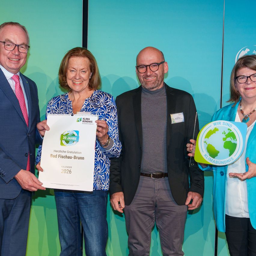 Vier Personen stehen vor einer blauen Wand und lächeln. Sie halten Auszeichnungen. Der Mann auf der linken Seite trägt einen Anzug und eine Krawatte. Die Frau neben ihm hält eine weiße Auszeichnung. Der Mann in der Mitte trägt eine Brille und einen dunklen Anzug. Die Frau auf der rechten Seite trägt eine Brille und eine blaue Jacke.