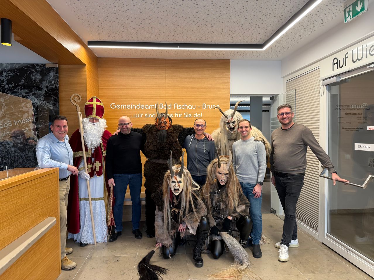 Gruppenfoto mit Maskottchen und Sankt Nikolaus im Büro. Sankt Nikolaus in rotem und weißem Gewand steht links. Vier Personen vor ihm tragen Masken und Kostüme.