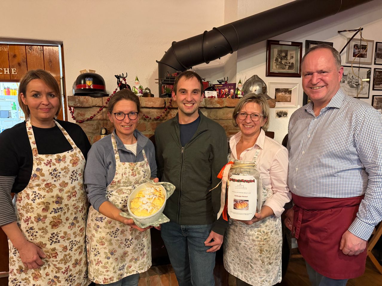 Fünf Menschen stehen lächelnd in einem Raum. Zwei Frauen in Schürzen halten Essen. Ein Mann in einer Jacke hält eine Glasflasche. Hinter ihnen ist ein Kamin mit Dekorationen.