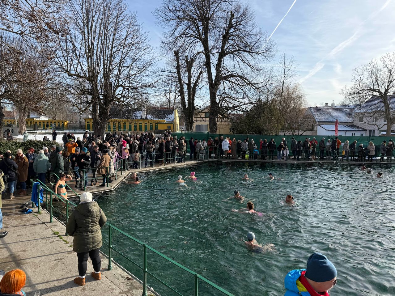 Eine Gruppe von Menschen schwimmt in einem Freibad, während viele andere am Zaun stehen, um zuzusehen. Der Pool ist von Bäumen umgeben, und im Hintergrund befindet sich ein Gebäude mit einem gelben Dach.