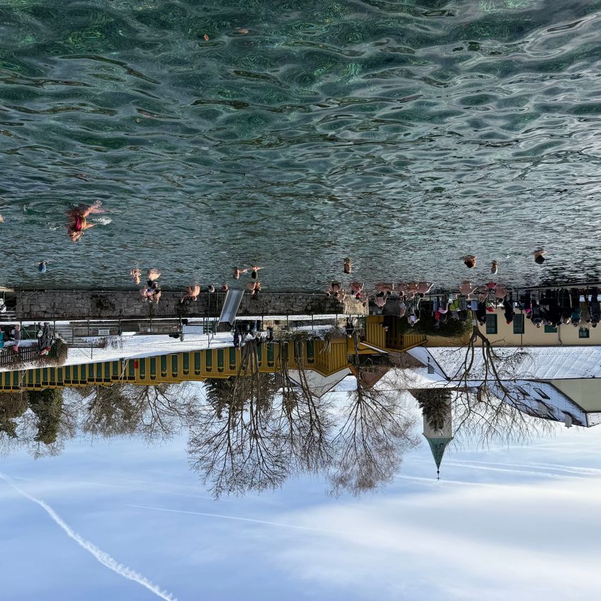 Eine Luftaufnahme von Menschen, die in einem See schwimmen, mit einer Brücke, Gebäuden und Bäumen, die sich im Wasser spiegeln.