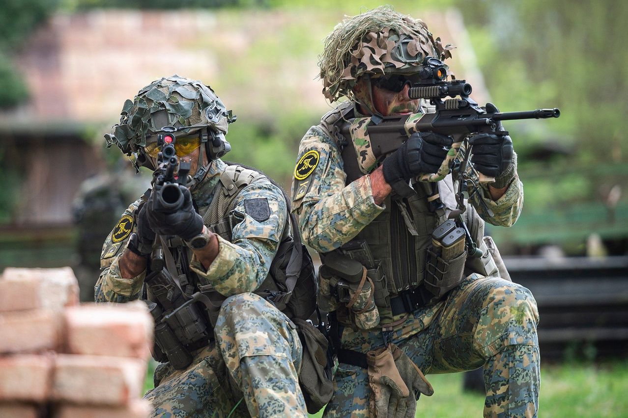 Zwei Soldaten in Tarnuniformen sind gekauert, einer hält eine Waffe und zielt, während der andere zur Seite schaut. Sie befinden sich in einem grasigen Bereich mit einem Ziegelstein im Vordergrund.