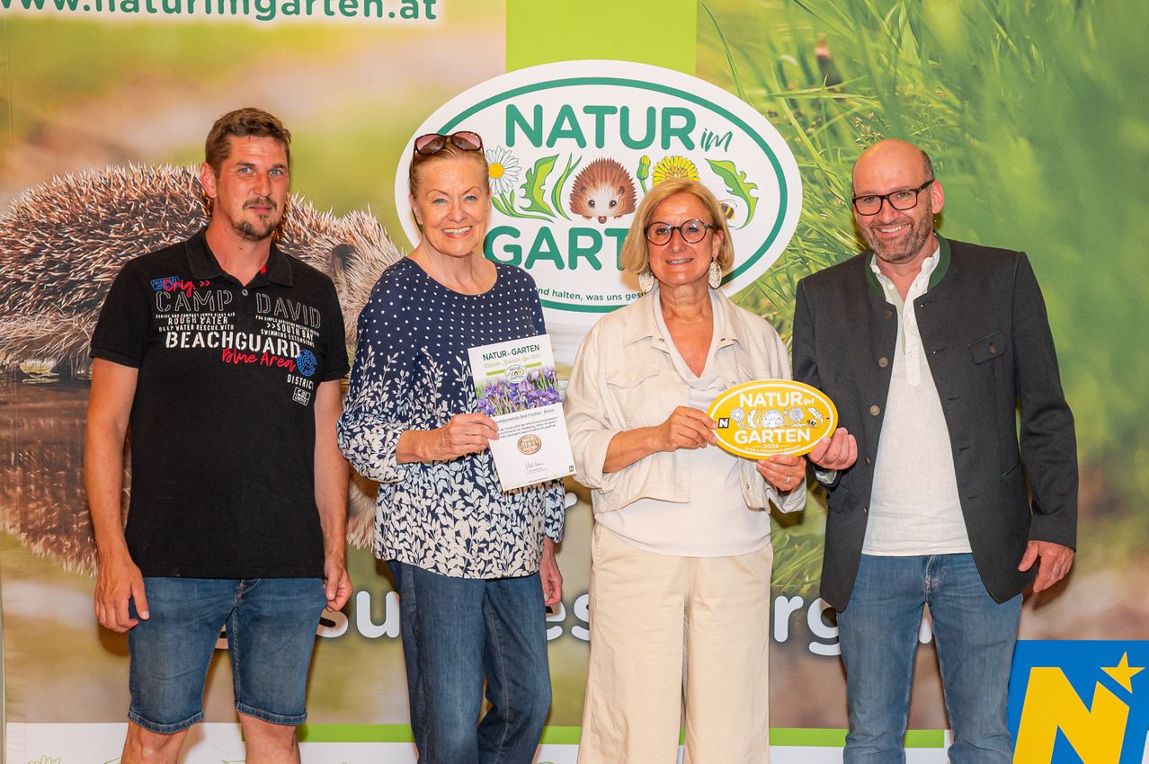 Vier Personen stehen vor einem Schild. Zwei Frauen halten ein Zertifikat und ein gelbes Schild, während ein Mann ein Zertifikat hält. Sie lächeln und scheinen für ein Foto zu posieren.