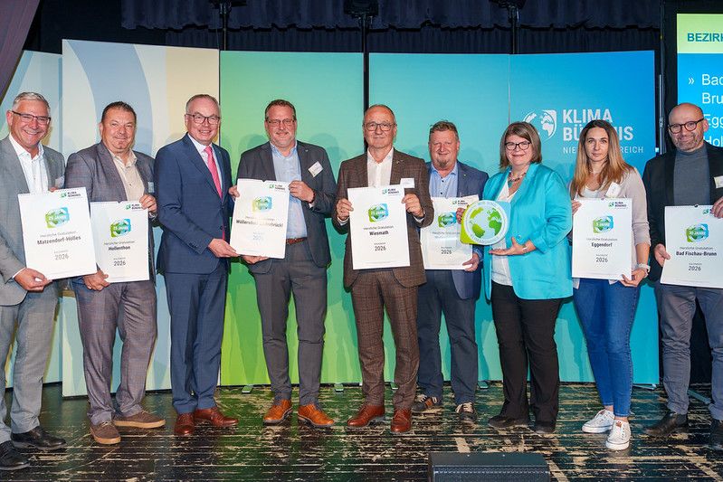 Nine individuals stand on a stage holding certificates, smiling for a photo. They wear formal attire, and a green backdrop with logos is behind them.