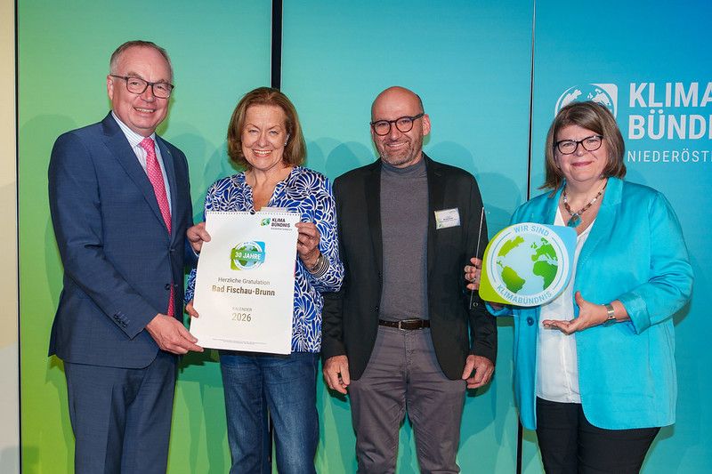 Four people stand in front of a green backdrop. They are holding certificates and smiling for a photo. The man on the left is in a blue suit. The woman next to him holds a certificate with the words 'Bad Fischau-Brunn 2026' on it. The man next to her wears glasses and a black suit. The woman on the right wears a blue jacket.