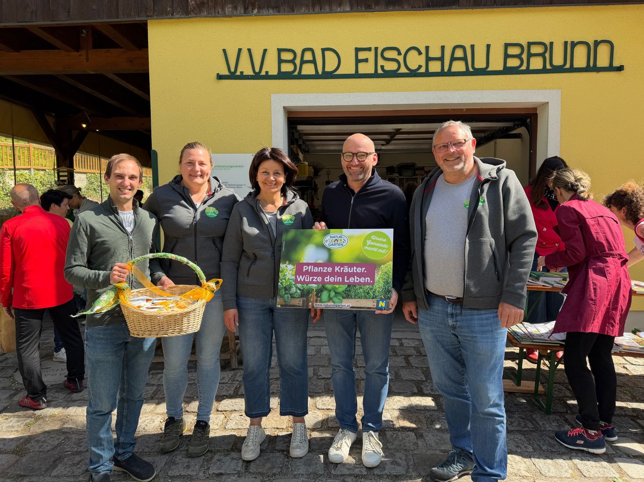 Five people stand outside a building with a yellow wall. They smile and pose for a photo. One person holds a sign and another a basket.