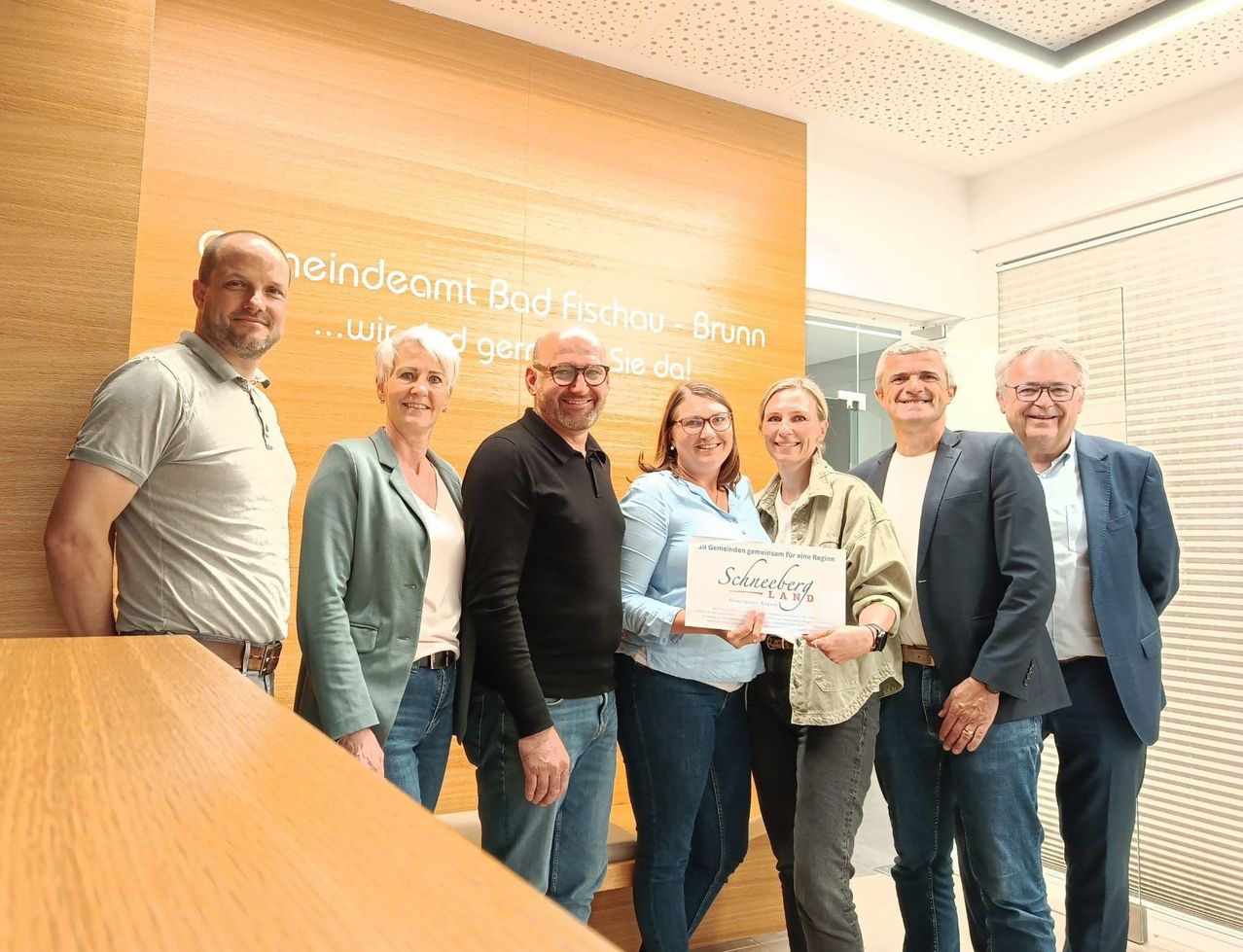 A group of people, including a woman holding a certificate, stand in front of a wooden wall with text. They seem to be posing for a photo.