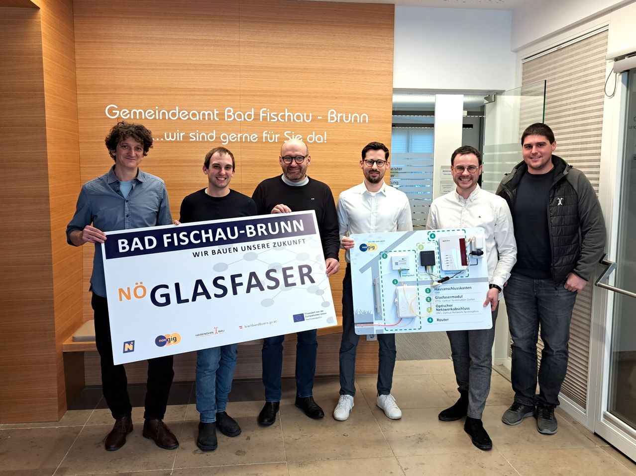 Fünf Männer stehen vor einer Holzwand und halten Schilder und ein Gerät in der Hand. Sie lächeln und scheinen eine neue Technologie zu bewerben. Das Schild zeigt 'Bad Fischau-Brunn' und 'NÖ Glasfaser.' Das Gerät ist ein Router.