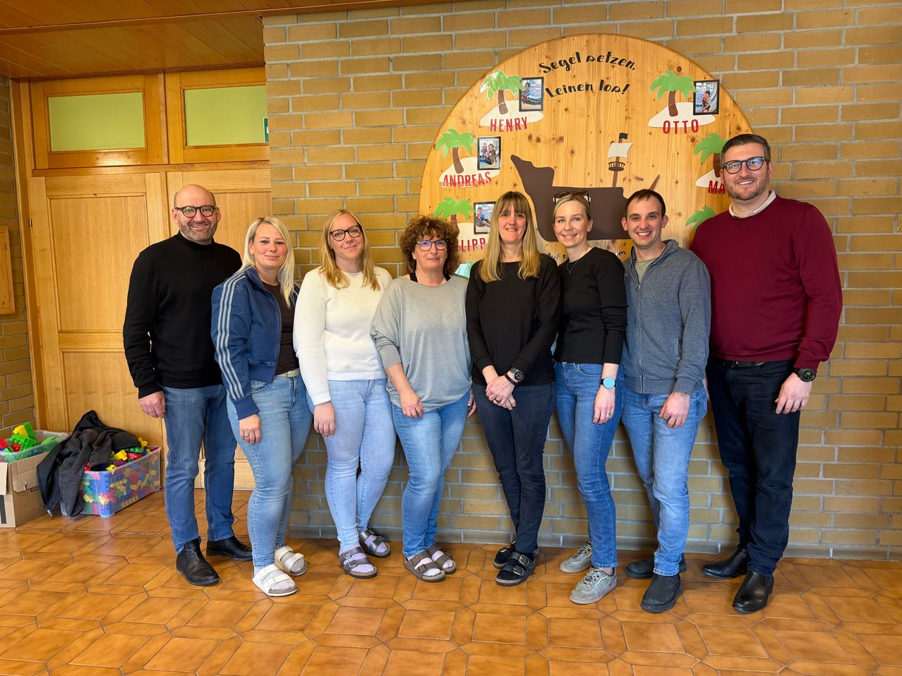 A group of adults, likely colleagues, are standing in front of a brick wall with a wooden board featuring a drawing of a sailboat and the text 'Segel pelzen. Leinen los!'. They are all smiling and seem to be posing for a photo.