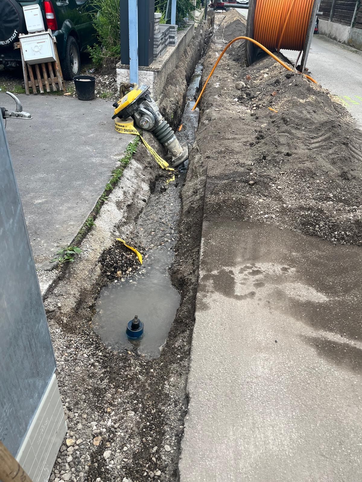 Ein offener Graben auf dem Bürgersteig mit einer Wasserleitung, einem gelben Warnband und einem schwarzen Container.