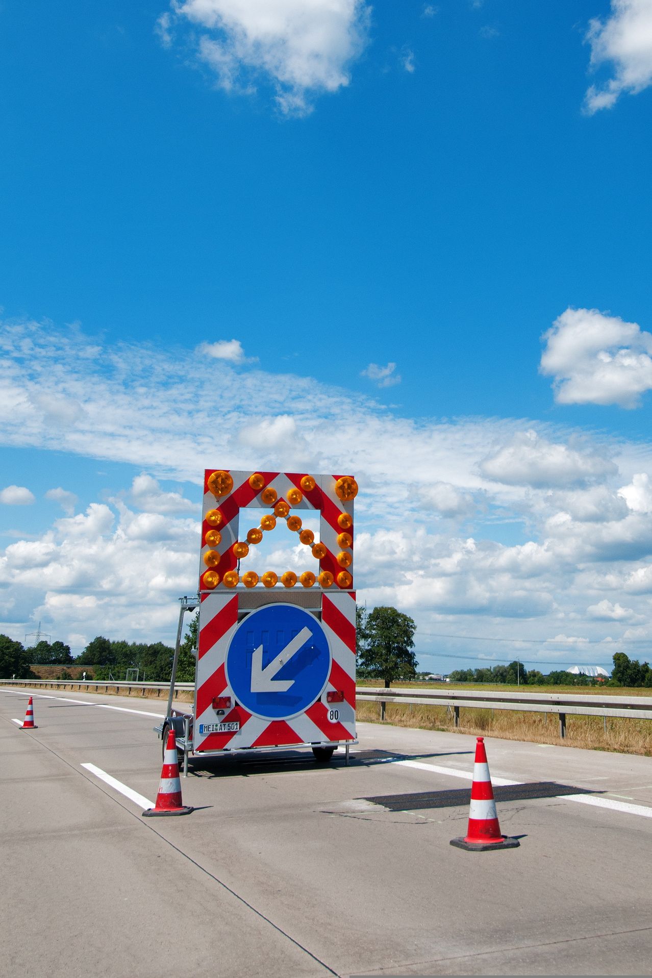 Eine Autobahn mit Verkehrskegeln und einem Schild, das eine Linkskurve anzeigt. Das Schild hat blinkende Lichter. Bäume und Gebäude sind in der Ferne unter einem bewölkten Himmel zu sehen.