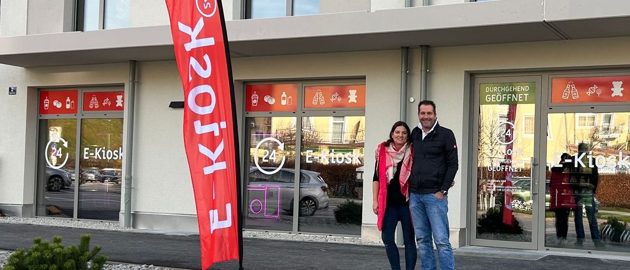 Zwei Personen stehen vor einem Gebäude mit einem roten Banner mit dem Wort 'e-Kiosk' darauf.