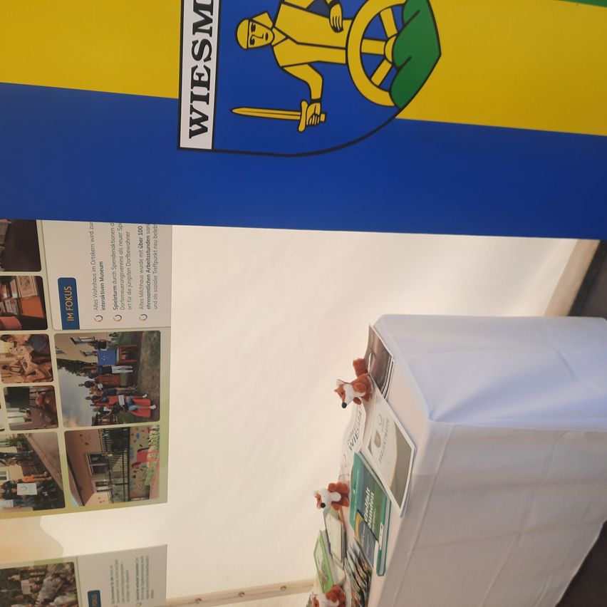 Ein weißer Banner mit blauen und gelben Streifen und einem Wappen ist über einem Tisch mit Broschüren und Spielzeug angebracht.