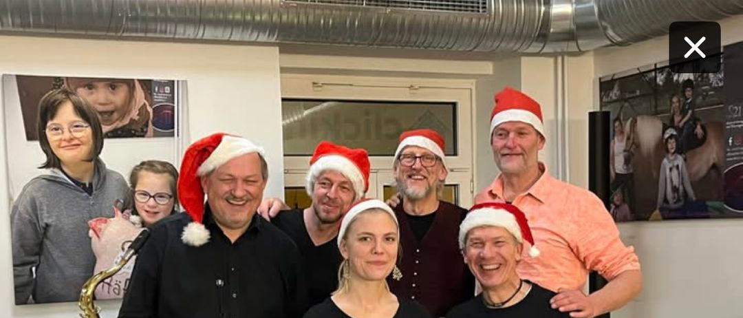 Eine Gruppe von Menschen in Weihnachtsmützen posiert in einem Raum. Eine Saxophon steht in der linken Ecke, und ein Fotorahmen ist an der Wand. Der Text lautet 'Save The Date 12 Dezember 2025 um 19 Uhr das Weihnachtskonzert der Haferband im Cafe Hafer'.