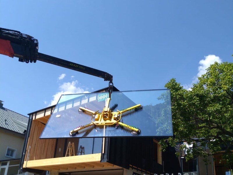 Ein Kran hebt ein großes Glaspanel von einer Baustelle unter einem klaren blauen Himmel mit verstreuten Wolken.