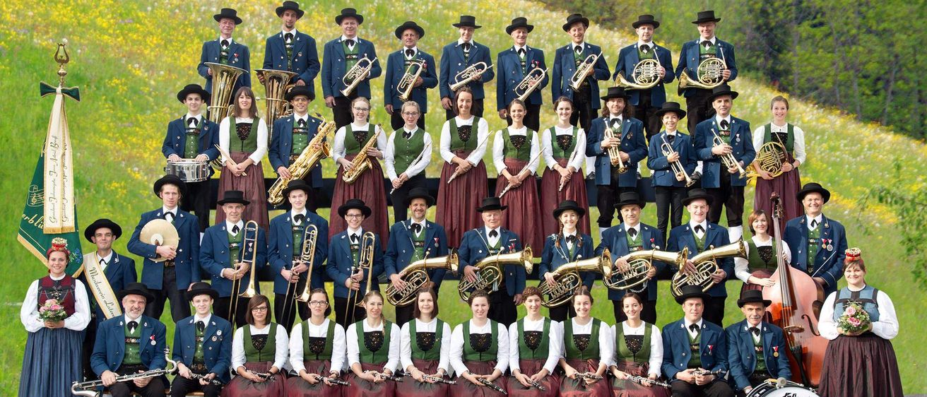 Eine große Gruppe von Musikern in traditioneller Kleidung posiert für ein Foto auf einer Wiese und hält verschiedene Instrumente wie Trompeten, Posaunen und Flöten. Sie sind von üppiger Grünfläche umgeben.