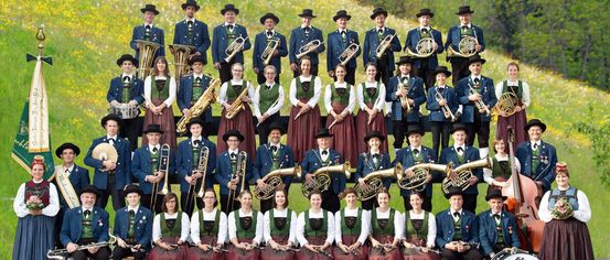 Eine große Gruppe von Musikern in traditioneller Kleidung posiert für ein Foto auf einer Wiese und hält verschiedene Instrumente wie Trompeten, Posaunen und Flöten. Sie sind von üppiger Grünfläche umgeben.