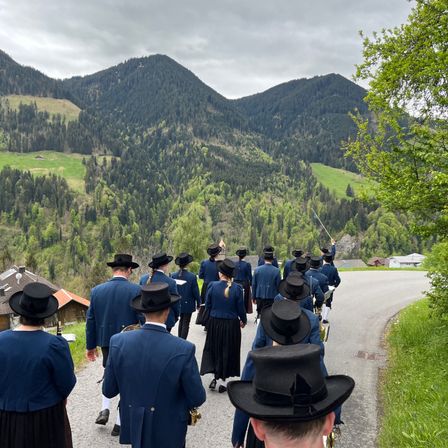 Eine Gruppe von Menschen in traditioneller Kleidung, die eine Straße entlangmarschiert, mit Bergen und Grün im Hintergrund.