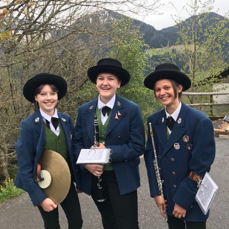 Drei junge Frauen in Uniformen, jede mit einem anderen Musikinstrument, lächeln und posieren für ein Foto vor einer Bergkulisse.