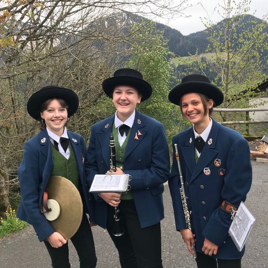 Drei junge Frauen in Uniformen, jede mit einem anderen Musikinstrument, lächeln und posieren für ein Foto vor einer Bergkulisse.