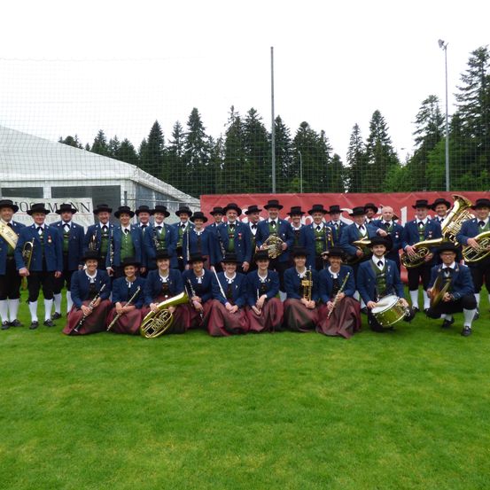 Eine Gruppe von Menschen in traditioneller Kleidung mit Musikinstrumenten, die für ein Foto auf einem Feld mit Bäumen und einem Zelt im Hintergrund posiert.