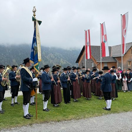 Eine Gruppe von Musikern in traditioneller Kleidung steht auf einem Rasenfeld und hält verschiedene Instrumente. Dahinter wehen Fahnen und ein Haus mit grauem Dach ist sichtbar. Im Hintergrund befinden sich Berge und ein bewölkter Himmel.