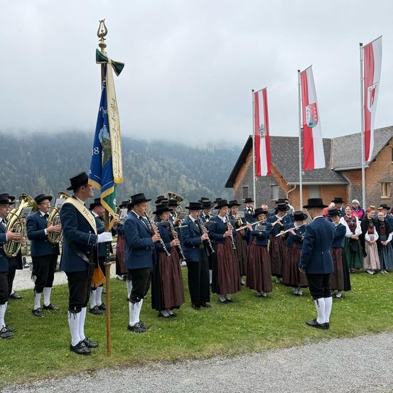 Eine Gruppe von Musikern in traditioneller Kleidung steht auf einem Rasenfeld und hält verschiedene Instrumente. Dahinter wehen Fahnen und ein Haus mit grauem Dach ist sichtbar. Im Hintergrund befinden sich Berge und ein bewölkter Himmel.