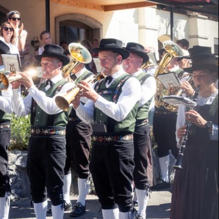 Eine Blaskapelle in traditioneller bayerischer Kleidung spielt im Freien, mit Musikern, die Trompeten, Posaunen und Klarinette spielen. Zuschauer schauen von hinten zu, einige klatschen.