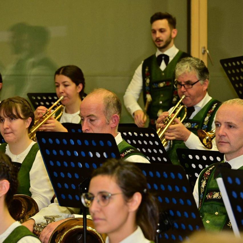 Eine Gruppe von Musikern in grünen und weißen Westen spielt Blechblasinstrumente in einem Raum. Sie schauen nach vorne und bereiten sich wahrscheinlich auf eine Aufführung vor.