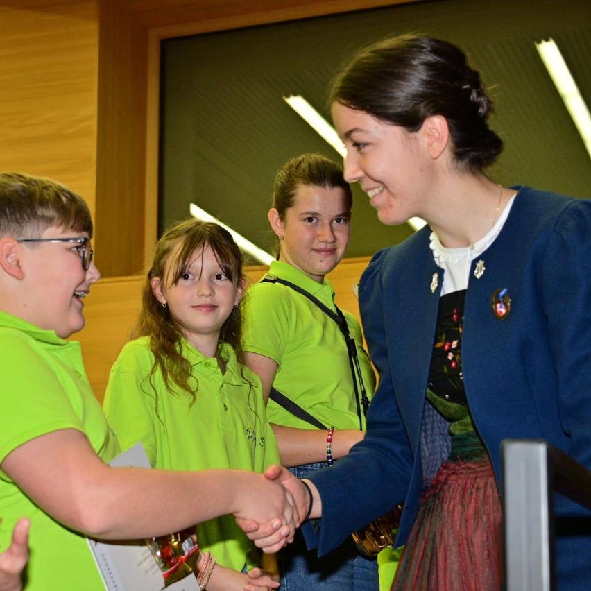Drei Kinder in grünen Shirts lächeln und schütteln die Hände einer Frau in einer blauen Jacke und mit einer Halskette. Hinter ihnen ist eine Wand mit einem Fenster und Lichtern zu sehen.