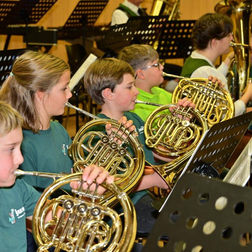 Eine Gruppe von Kindern, gekleidet in grünen T-Shirts, spielt französische Hörner und Trompeten in einem Klassenzimmer. Notenständer mit Notenblättern sind vor ihnen sichtbar.