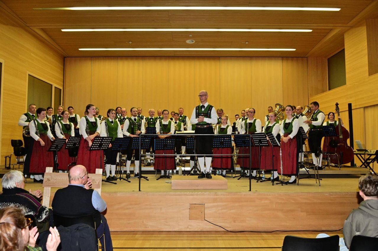 Ein Chor, gekleidet in traditioneller grüner und roter Kleidung, tritt auf einer Bühne mit Musikinstrumenten auf. Ein Mann steht in der Mitte und trägt eine Brille. Das Publikum sitzt vorne.