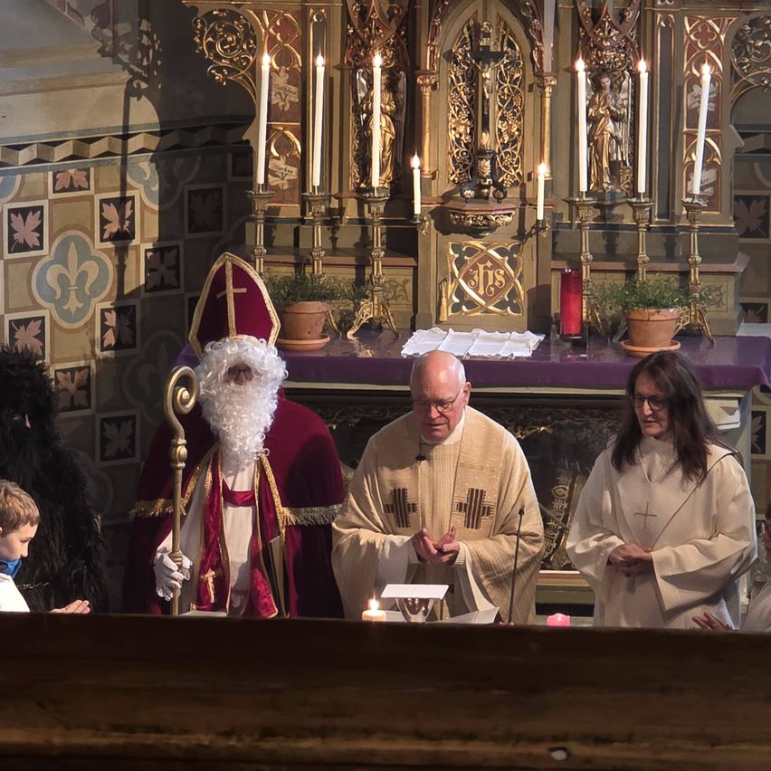 A priest and two assistants stand before a decorated altar with burning candles. A man dressed as Santa Claus holds a staff and stands behind them. A boy watches from below.