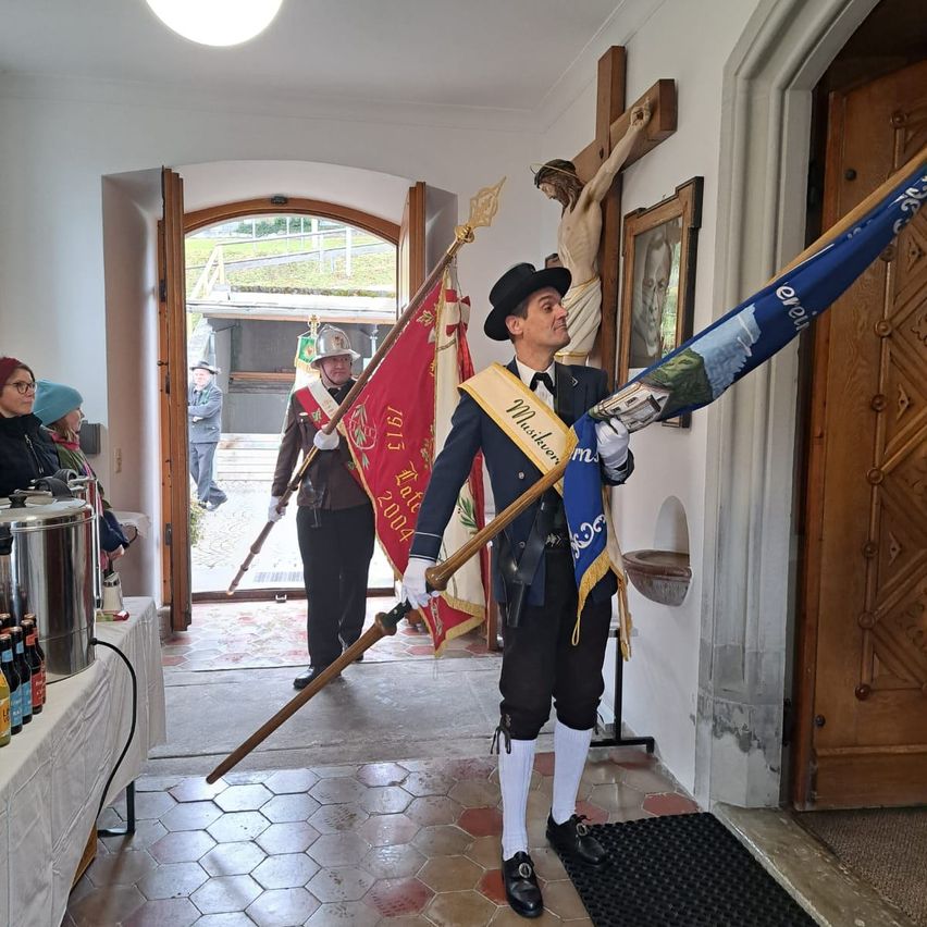 A man in traditional attire holding a flag and a pole stands inside a church, while two men in uniforms and hats stand behind him. One man holds a flag and a pole. A crucifix is mounted on the wall.