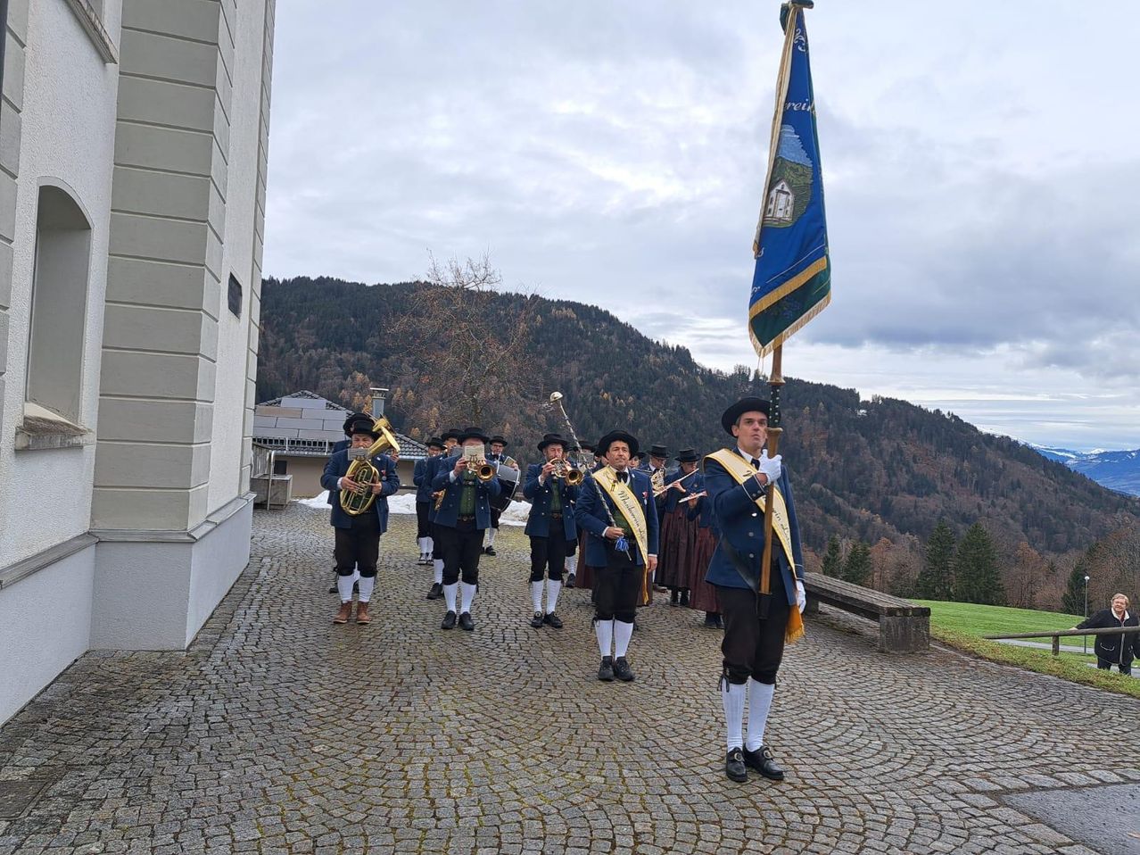 A brass band in traditional attire is marching in a procession with a flag, against a backdrop of mountains.