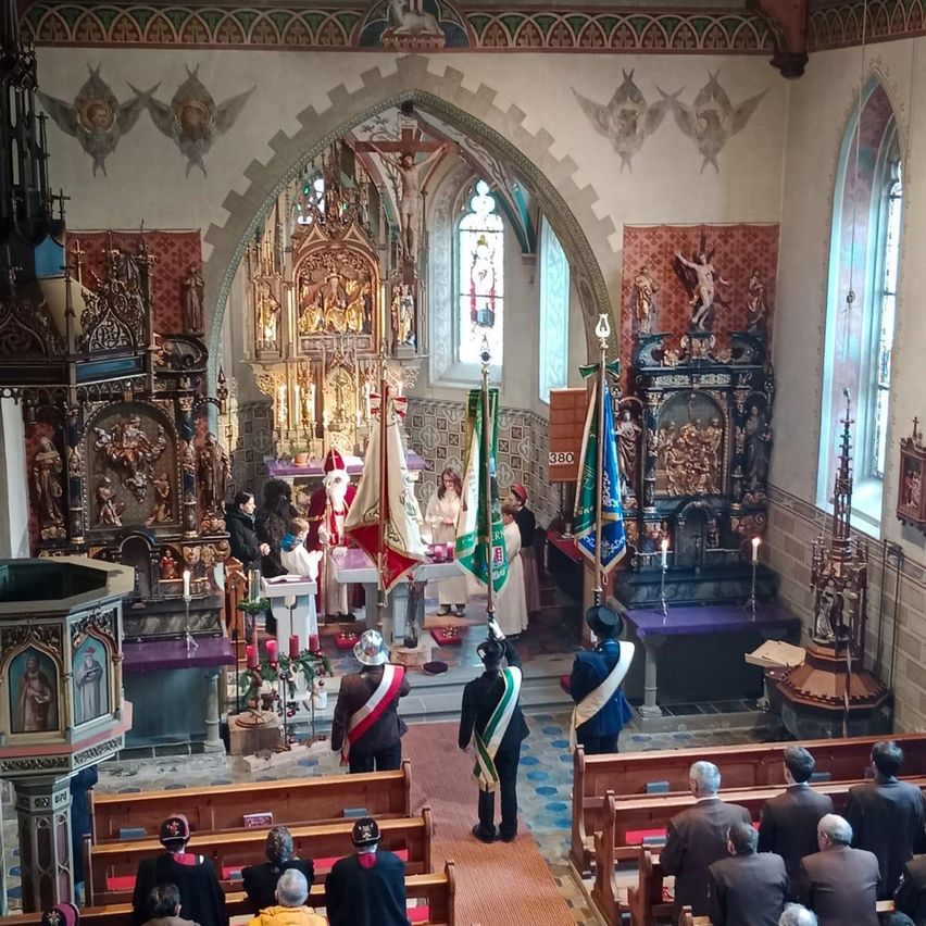 A solemn ceremony takes place in a grand church. People stand near the altar, while others sit in pews. The church is adorned with intricate carvings, candles, and flags. An altar with a statue of Jesus is in the center.