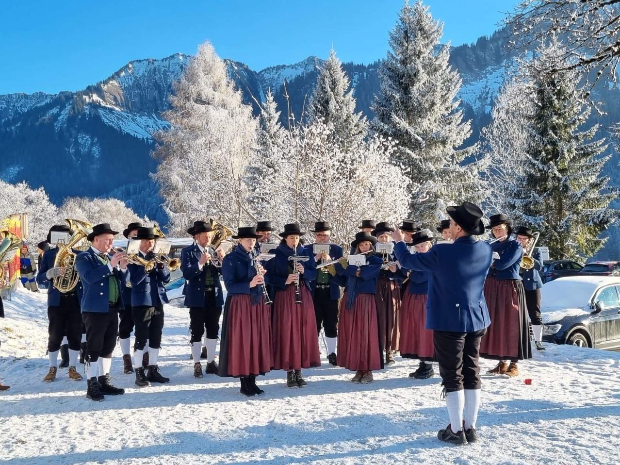 Eine Gruppe Musiker in traditioneller Winterkleidung tritt auf einem verschneiten Feld auf, mit verschneiten Bergen im Hintergrund.