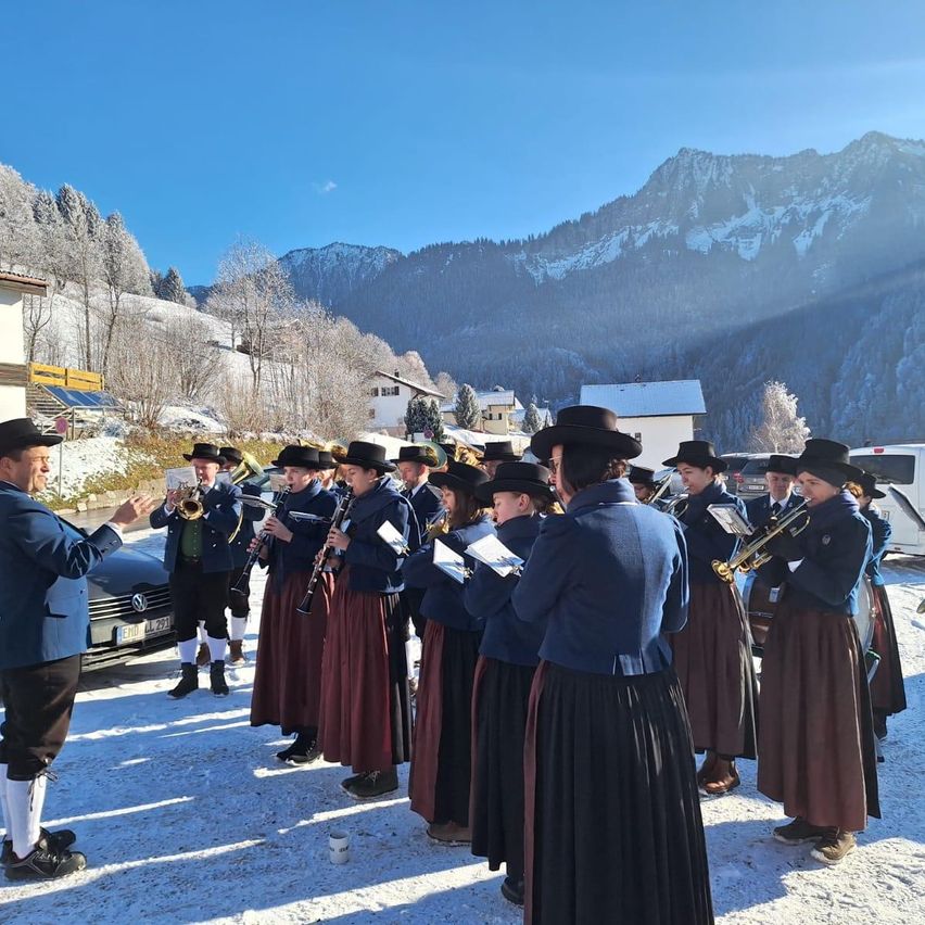 Ein Blasorchester in traditioneller Kleidung spielt in einer verschneiten alpinen Umgebung, mit Bergen im Hintergrund und einem Mann, der sie anführt.