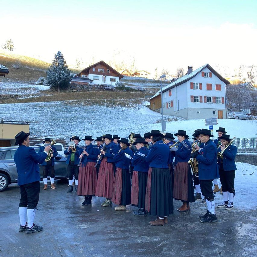 Ein Marschkapelle in traditioneller Kleidung tritt in einem verschneiten Gebirgsgebiet auf, mit Häusern und einem Auto im Hintergrund.