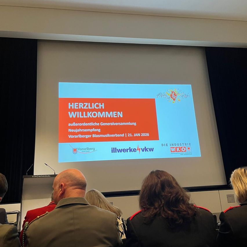 Eine Gruppe von Personen in formeller Kleidung sitzt vor einer Projektionsleinwand, die eine Präsentation zeigt. Die Folie lautet Herzlich Willkommen mit rotem Rand. Datum 21. Jan 2026 und Logo sichtbar.