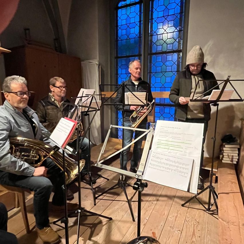 Four men are preparing to perform with their brass instruments. They are standing in front of music stands in a room with wooden floors and stained glass windows.