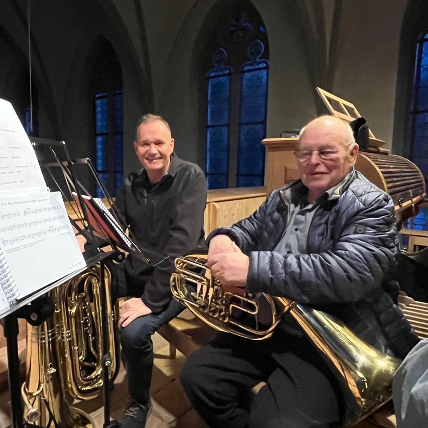Two men are seated in a church, one playing a tuba and the other looking at sheet music. A wooden organ is behind them.
