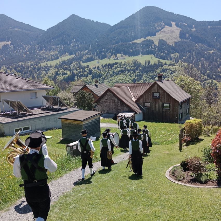 A marching band of men and women in traditional attire walks down a grassy path in a village, with houses and mountains in the background.