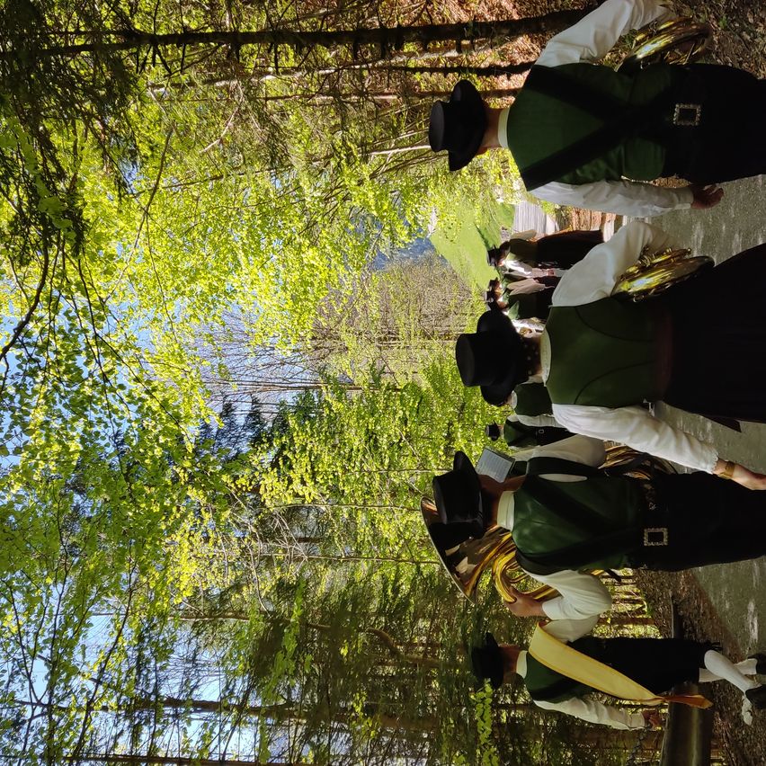A group of musicians in green vests and black pants play instruments on a path surrounded by green trees.